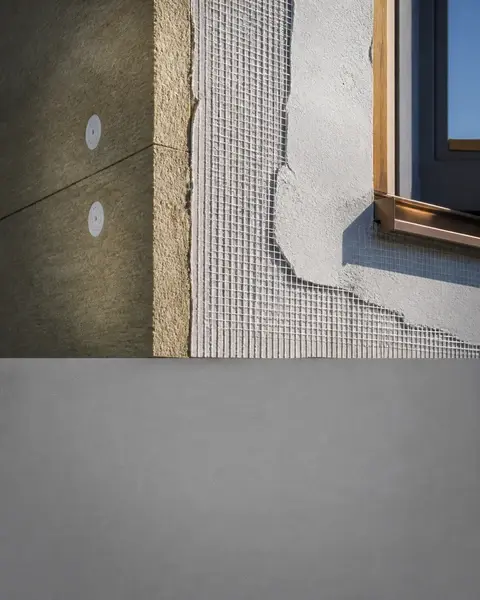Präziser Schichtaufbau einer Fassadendämmung im streifenden Tageslicht: Mineralwollplatten, Armierung und halb aufgezogener Oberputz