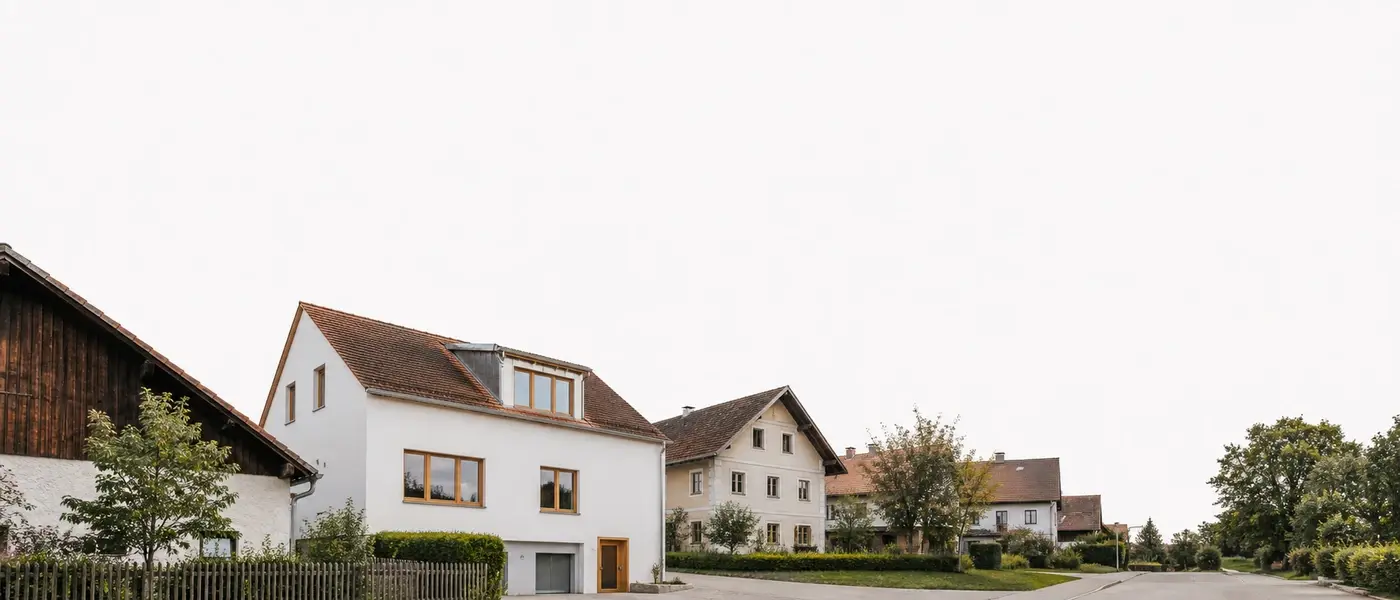 Ruhige bayerische Wohnstraße bei bedecktem Tageslicht, ein frisch modernisiertes Haus mit weißem Putz zwischen älteren Nachbarhäusern